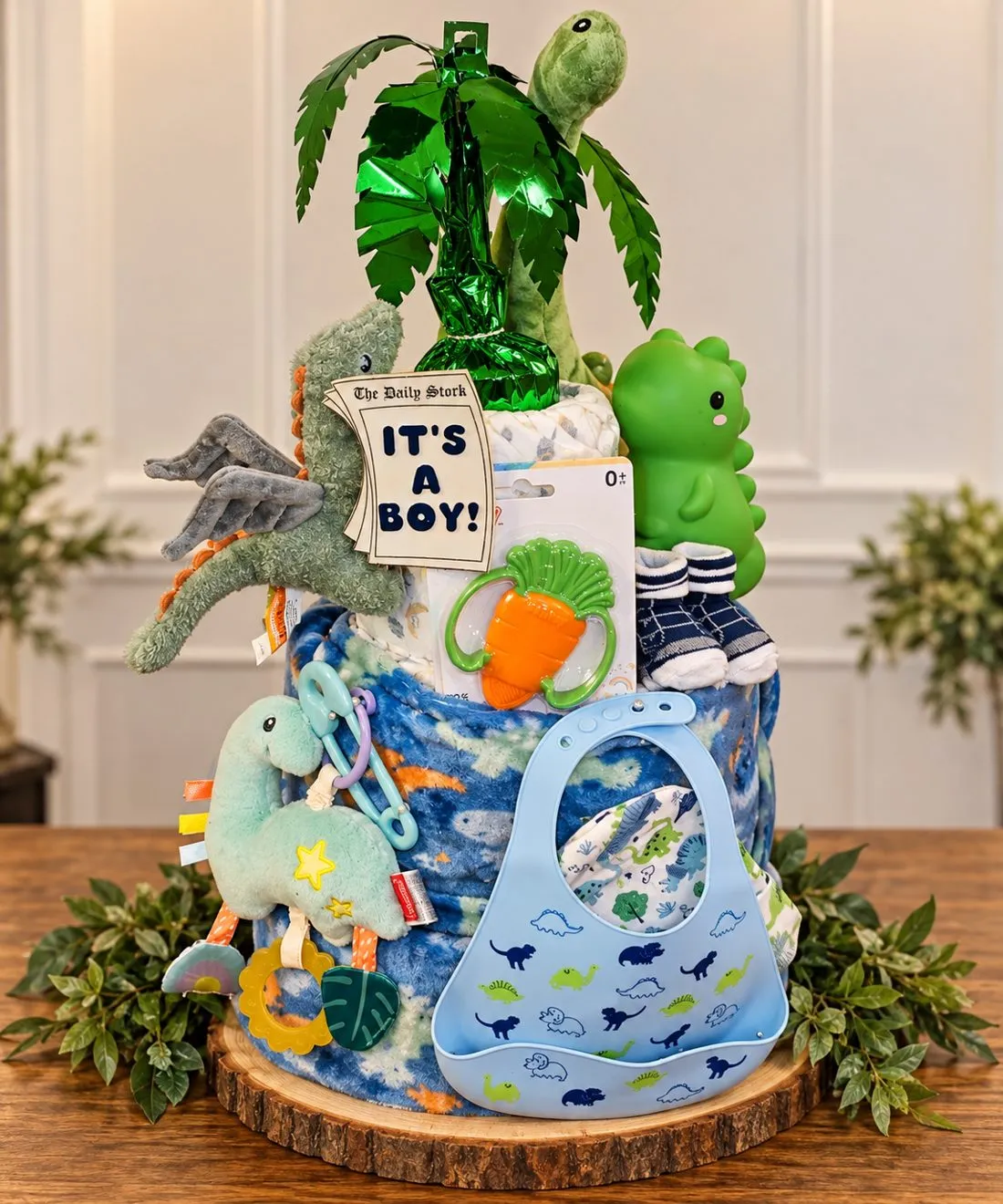 Diaper cake decorated with dinosaur toys, a palm leaf and a blue bib on a wooden slice